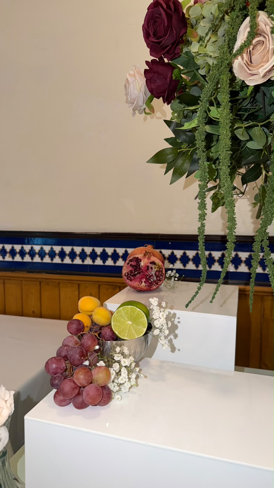 Pomegranate, grapes and apricots on white plinths beneath hanging roses